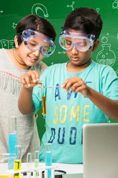 2 kleine indische Kinder beim naturwissenschaftlichen Experiment oder Chemie-Experiment im Klassenzimmer mit Schutzbrille auf grünem Kreidetafelhintergrund mit darüber gezogenen naturwissenschaftlichen Kritzeleien — Stockfoto