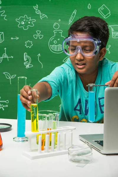 Kinder und Naturwissenschaftliches Konzept - niedlicher indischer kleiner Junge beschäftigt mit naturwissenschaftlichen oder chemischen Experimenten mit Reagenzglas und Kolben mit Sicherheitsaugglas über grünem Kreidetafelhintergrund mit gezeichneten Naturwissenschaftskritzeleien — Stockfoto