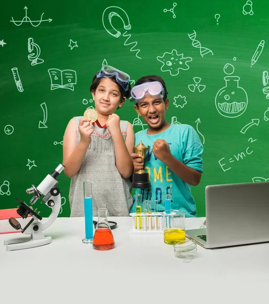 2 crianças indianas fazendo experimento de ciência ou experimento de química em sala de aula com óculos de segurança em pé sobre fundo de quadro-negro verde com doodles científicos desenhados sobre ele — Fotografia de Stock