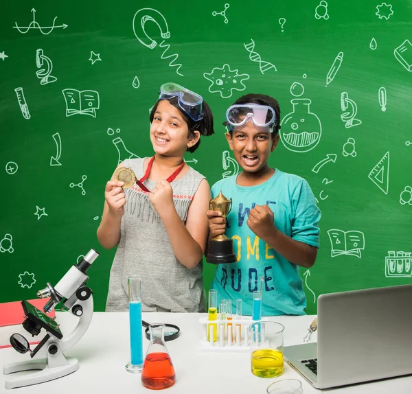 2 crianças indianas fazendo experimento de ciência ou experimento de química em sala de aula com óculos de segurança em pé sobre fundo de quadro-negro verde com doodles científicos desenhados sobre ele — Fotografia de Stock