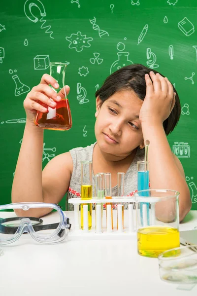Kids and Science concept - Cute Indian little girl busy doing science or chemistry experiment with test tube and flask with safety eye glass over green chalkboard background with science doodles drawn
