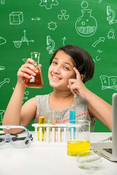 Kids and Science concept - Cute Indian little girl busy doing science or chemistry experiment with test tube and flask with safety eye glass over green chalkboard background with science doodles drawn