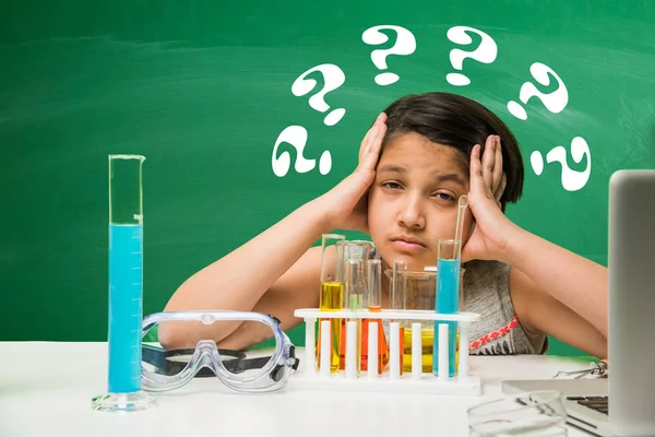 Kids and Science concept - Cute Indian little girl worried or confused or sad while doing science or chemistry experiment with test tube / flask over green chalkboard background & science doodles