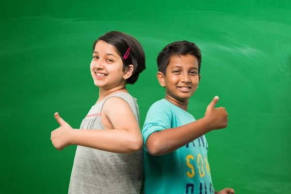 Cute indian boy and girl or school kids standing in front of empty green chalkboard background showing success or victory symbol with thumbs up — Stock Photo, Image