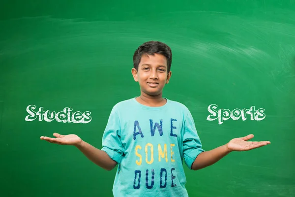Indian school kid or boy standing in front of green chalkboard with both hands stretched, presenting some concept with copy space — Stock Photo, Image