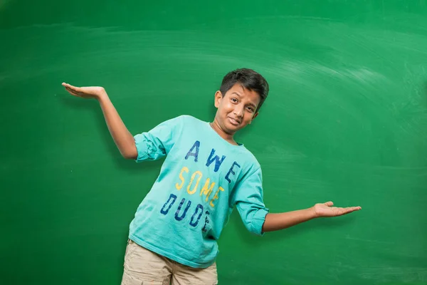 Indian school kid or boy standing in front of green chalkboard with both hands stretched, presenting some concept with copy space — Stock Photo, Image