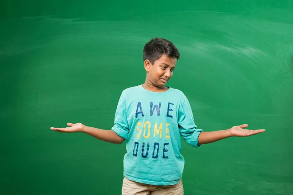 Indian school kid or boy standing in front of green chalkboard with both hands stretched, presenting some concept with copy space — Stock Photo, Image