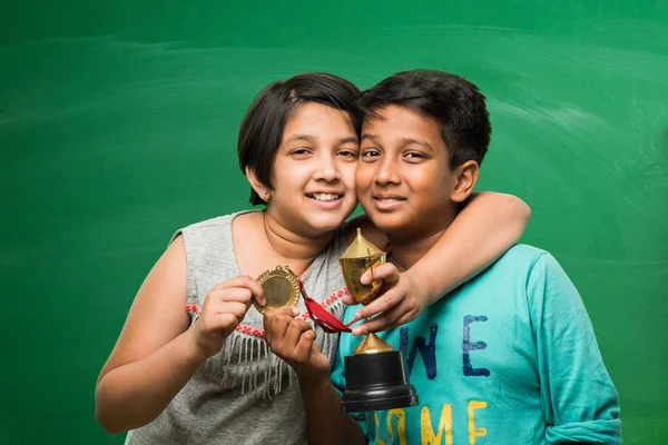 Indian smart girl and boy or school kids holding gold medal and trophy cup over green chalkboard background — Stock Photo, Image