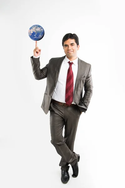Foto completa del joven empresario indio mirando la cámara mientras equilibra el globo azul o el modelo de tierra en el dedo índice y lleva un atuendo corporativo completo como traje y corbata, aislado sobre fondo blanco — Foto de Stock