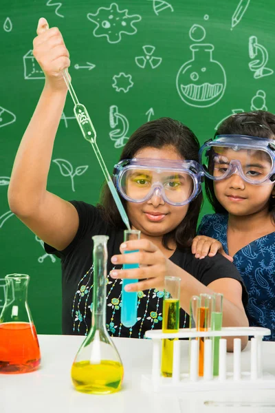 2 süße kleine indische Mädchen machen naturwissenschaftliche Experimente oder Projekte in einem Klassenzimmer mit grüner Kreidetafel mit naturwissenschaftlichen Kritzeleien — Stockfoto