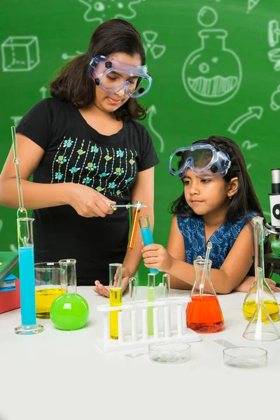 2 süße kleine indische Mädchen machen naturwissenschaftliche Experimente oder Projekte in einem Klassenzimmer mit grüner Kreidetafel mit naturwissenschaftlichen Kritzeleien — Stockfoto