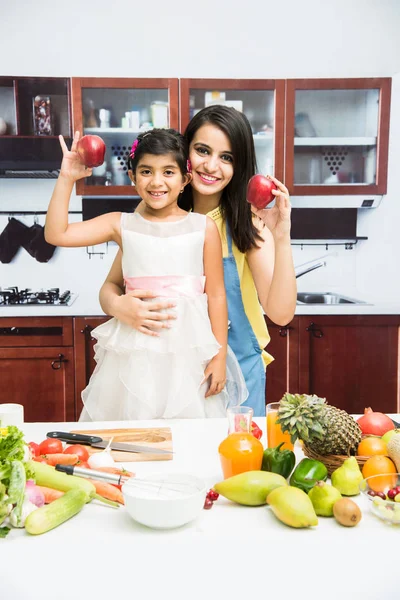 Pretty Indian young lady or mother with cute girl child or daughter in kitchen having fun time with table full of fresh vegetables and fruits