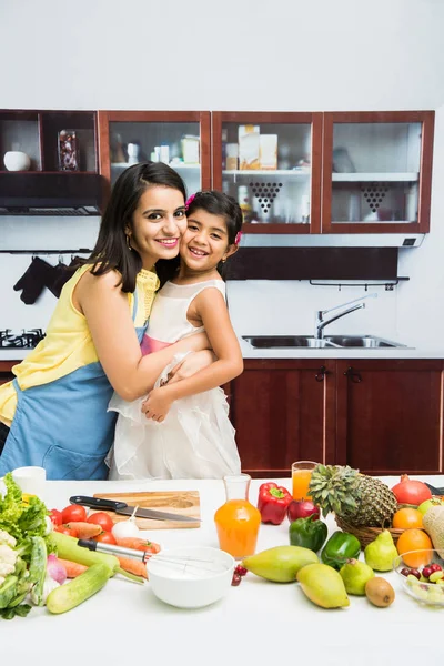 Pretty Indian young lady or mother with cute girl child or daughter in kitchen having fun time with table full of fresh vegetables and fruits