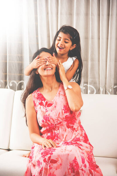 playful happy Indian little girl covering her mother's eyes and giving her surprise while sitting on white sofa in drawing room