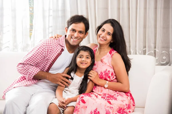 Family time - Candid Portrait of smart indian family of three while sitting on sofa. Indian or asian family group photo. Cute girl child sitting between mother and father on sofa for portrait photo — Stock Photo, Image
