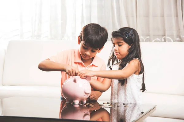 Indian small kids with piggy bank, asian little brother and sister putting coins inside piggy bank planning finances — Stock Photo, Image