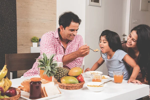 An attractive happy, smiling Asian Indian family of mother, father and daughter. Father feeding cereal to daughter with spoon at dining table. Indians eating breakfast, lunch / dinner. Selective focus — Stock Photo, Image