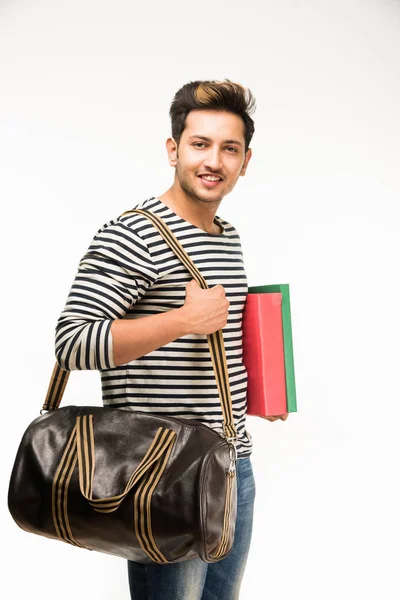 Bonito e jovem indiano estudante universitário masculino carregando saco em fundo branco enquanto segurando livros da faculdade, laptop ou telefone inteligente — Fotografia de Stock