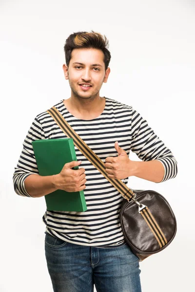 Bonito e jovem indiano estudante universitário masculino carregando saco em fundo branco enquanto segurando livros da faculdade, laptop ou telefone inteligente — Fotografia de Stock