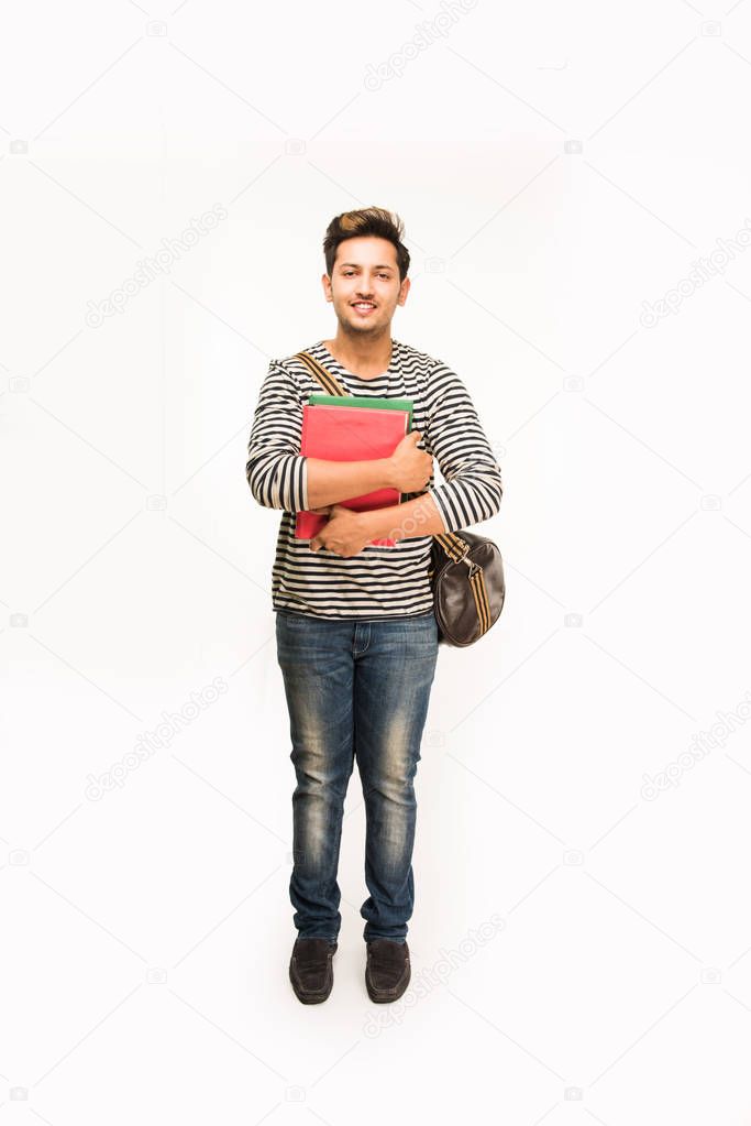 Handsome and young indian Male college student carrying bag on white background while holding college books, laptop or smart phone