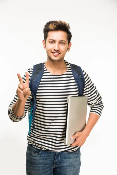 Guapo y joven indio estudiante universitario masculino que lleva el bolso en un fondo blanco mientras sostiene libros universitarios, computadora portátil o teléfono inteligente — Foto de Stock