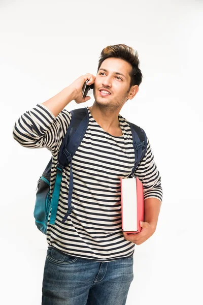 Bonito e jovem indiano estudante universitário masculino carregando saco em fundo branco enquanto segurando livros da faculdade, laptop ou telefone inteligente — Fotografia de Stock