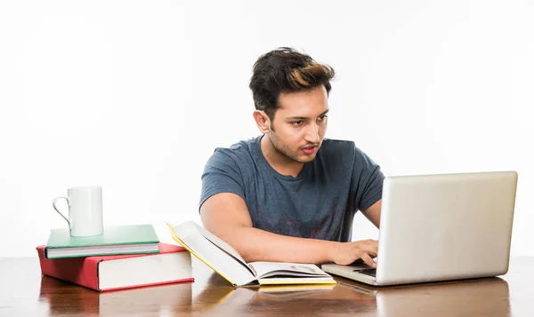 Beau garçon indien ou étudiant étudiant étudiant sur la table d'étude avec pile de livres, ordinateur portable et tasse à café. Sourire ou penser ou s'inquiéter ou montrer les pouces vers le haut ou en utilisant un smartphone — Photo