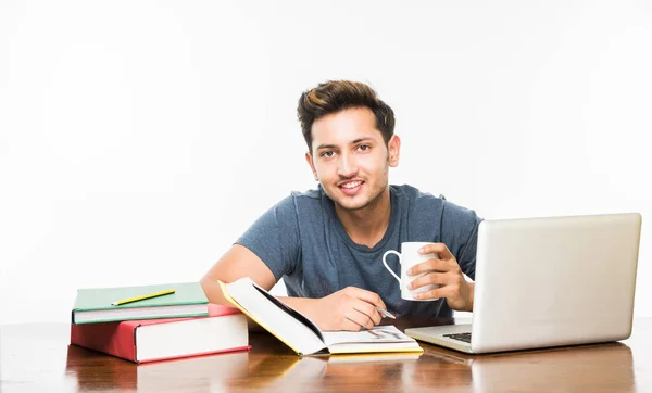 Vacker indisk pojke eller manlig collegestudent som studerar på studie bord med högen av böcker, bärbar dator och kaffe mugg. Ler eller tänkande eller orolig eller visar tummen eller använder smartphone — Stockfoto