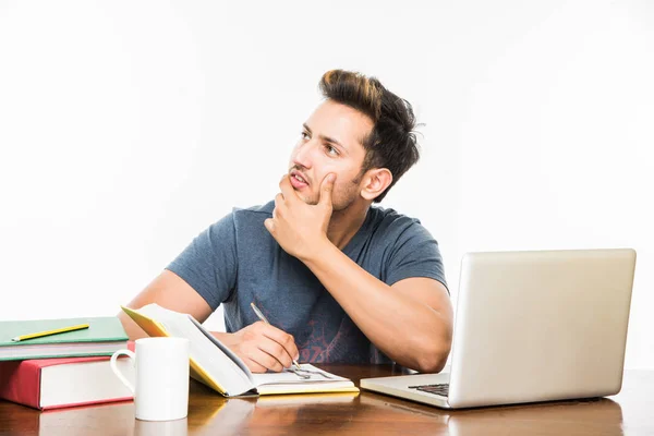 Beau garçon indien ou étudiant étudiant étudiant sur la table d'étude avec pile de livres, ordinateur portable et tasse à café. Sourire ou penser ou s'inquiéter ou montrer les pouces vers le haut ou en utilisant un smartphone — Photo