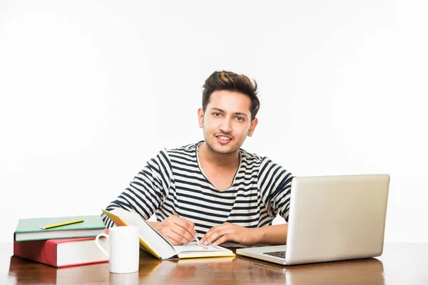 Beau garçon indien ou étudiant étudiant étudiant sur la table d'étude avec pile de livres, ordinateur portable et tasse à café. Sourire ou penser ou s'inquiéter ou montrer les pouces vers le haut ou en utilisant un smartphone — Photo