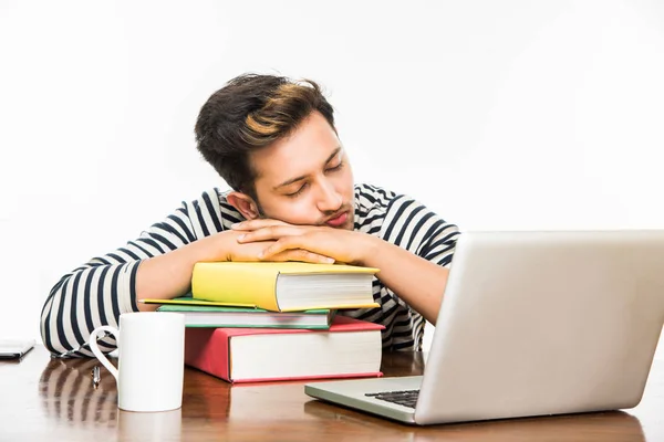 Handsome Indian boy or male college student studying on study table with pile of books, laptop computer and coffee mug. Smiling or thinking or worried or showing thumbs up or using smartphone — Stock Photo, Image