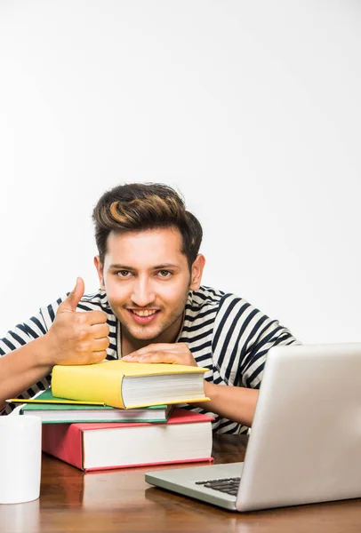 Guapo niño indio o estudiante universitario masculino que estudia en la mesa de estudio con pila de libros, computadora portátil y taza de café. Sonriendo o pensando o preocupado o mostrando los pulgares hacia arriba o usando el teléfono inteligente — Foto de Stock