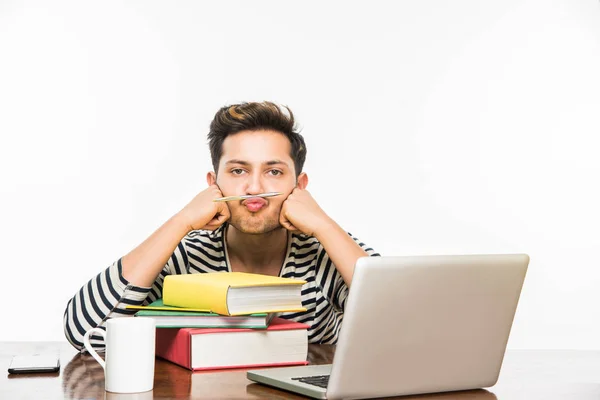 Beau garçon indien ou étudiant étudiant étudiant sur la table d'étude avec pile de livres, ordinateur portable et tasse à café. Sourire ou penser ou s'inquiéter ou montrer les pouces vers le haut ou en utilisant un smartphone — Photo