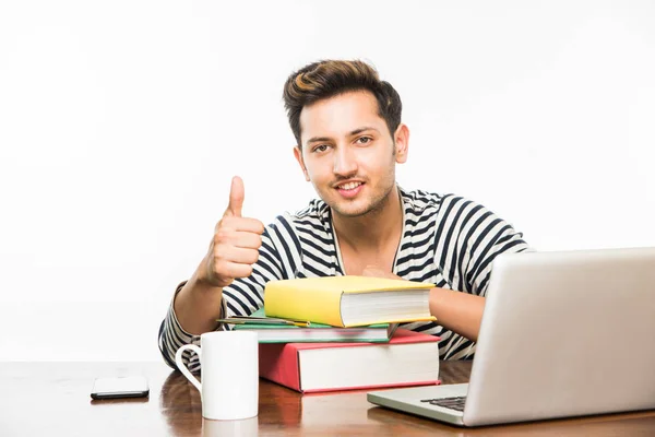 ハンサムなインド男の子または男性の大学生は勉強の本の杭の研究テーブルの上のラップトップ コンピューターとコーヒーのマグカップ。笑顔や思考または心配または親指を現して、スマート フォンを使用して — ストック写真