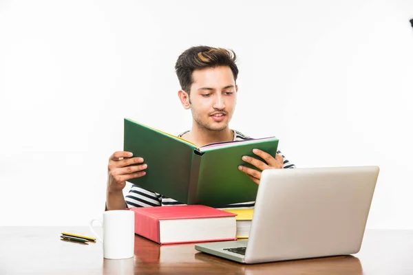 Beau garçon indien ou étudiant étudiant étudiant sur la table d'étude avec pile de livres, ordinateur portable et tasse à café. Sourire ou penser ou s'inquiéter ou montrer les pouces vers le haut ou en utilisant un smartphone — Photo