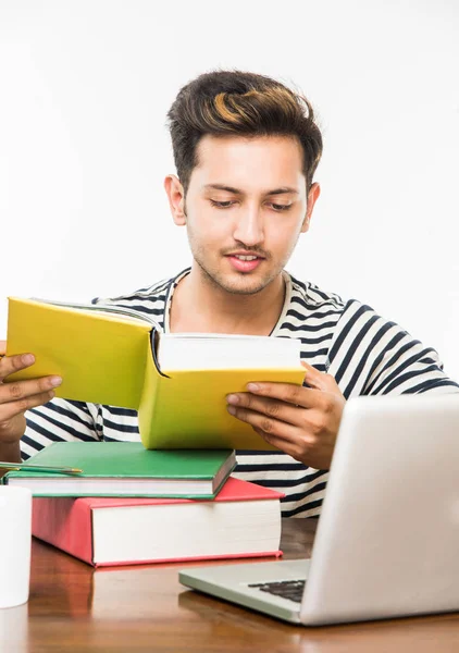 Menino indiano bonito ou estudante universitário masculino estudando na mesa de estudo com pilha de livros, computador portátil e caneca de café. Sorrindo ou pensando ou preocupado ou mostrando polegares para cima ou usando smartphone — Fotografia de Stock