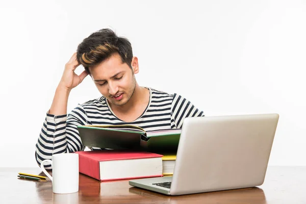 Beau garçon indien ou étudiant étudiant étudiant sur la table d'étude avec pile de livres, ordinateur portable et tasse à café. Sourire ou penser ou s'inquiéter ou montrer les pouces vers le haut ou en utilisant un smartphone — Photo