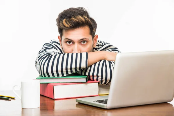 ハンサムなインド男の子または男性の大学生は勉強の本の杭の研究テーブルの上のラップトップ コンピューターとコーヒーのマグカップ。笑顔や思考または心配または親指を現して、スマート フォンを使用して — ストック写真