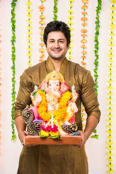 Indian handsome man in ethnic wear holding a Ganesh idol, welcoming God on Ganesh Chaturthi / festival at home with happy expressions