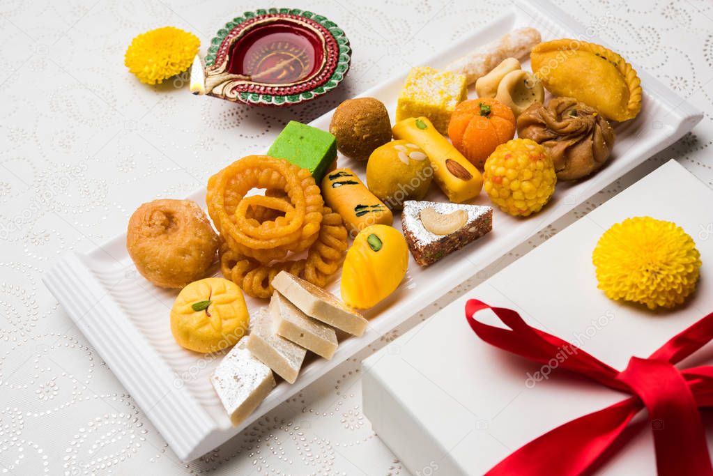 stock photo of Indian sweet or mithai and oil lamp or diya with gift box and flowers on decorative or colourful background, selective focus