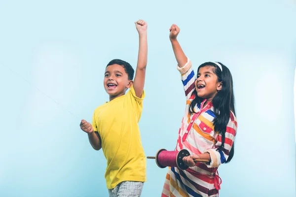 Drachen oder Patang fliegen in Indien, zwei süße kleine indische Kinder genießen Drachenfliegen in makar sankranti Festival, stehen mit Chakri oder Holzspindel und halten Faden in Aufregung auf blauem Hintergrund — Stockfoto
