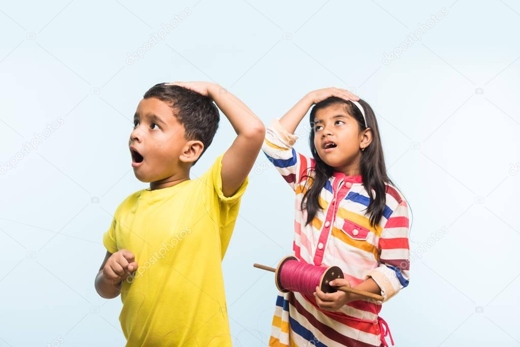 Kite or Patang flying in India, two cute little indian kids enjoying Kite flying in Makar sankranti festival, standing with chakri or wooden spindal and holding thread in excitement on blue background
