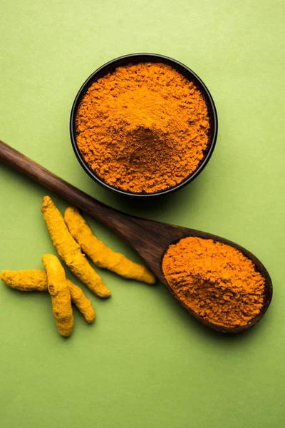 Turmeric powder in ceramic bowl with raw dried turmeric over plain background