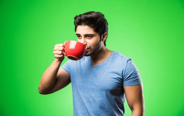 Indian man with tea / coffee cup or mug. Drinking, presenting or holding while standing isolated over green background