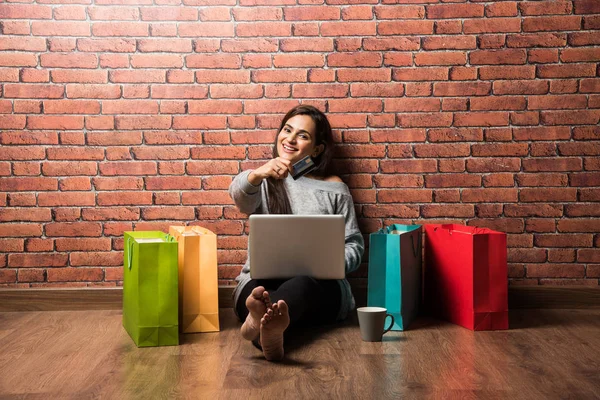 Indian girl shopping with debit / Credit card and laptop while sitting over wooden floor against red brick wall