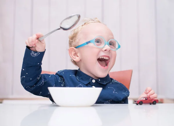 Criança usando óculos sentados à mesa e segurando uma colher em suas mãos — Fotografia de Stock