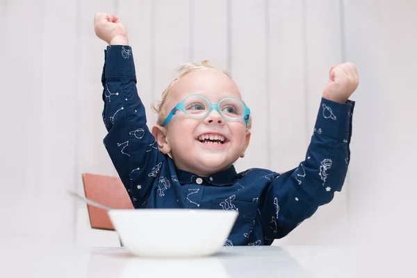 Criança usando óculos sentados à mesa e segurando uma colher em suas mãos — Fotografia de Stock
