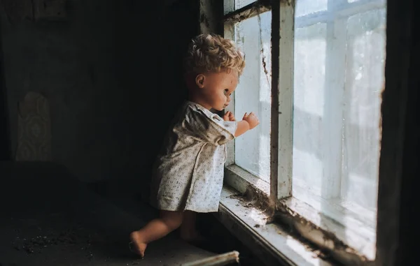 old doll stands by the window and looks out the window. The concept of loneliness, sadness, poverty.