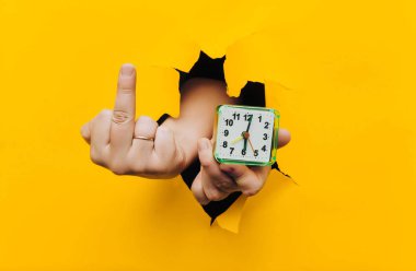 Bir erkek elinde yeşil alarmlı saat, ikincisinde de orta parmak var. Zaman kavramı ve erken kalkma ihtiyacı. Geçmişten nefret. Sarı arkaplan, yırtık delik, kopyalama alanı.