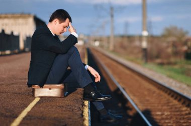 Üzgün genç bir işadamı, tren yolu geçmişinde bir bavulun üzerinde bir platformda oturuyor. İşsizlik, kriz, yaptırımlar. Rus işi.
